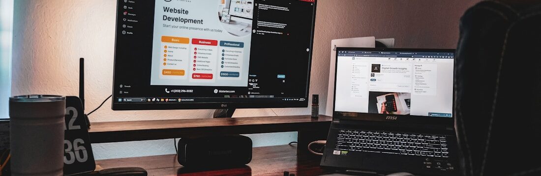 A dimly lit workspace featuring a desktop monitor displaying a website development page and a laptop showing a digital growth insights page. The setup includes a keyboard, mouse, two cups, a pen holder, and a clock displaying '02 36'. The atmosphere suggests a cozy, focused work environment.